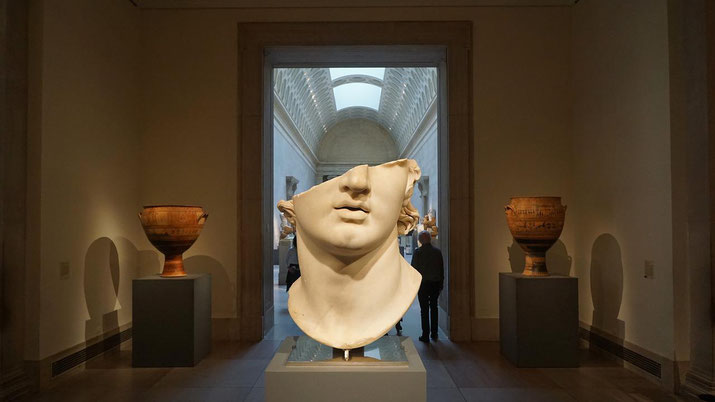 Large marble head with missing upper features is displayed between ancient vases in a museum gallery with arched ceilings.