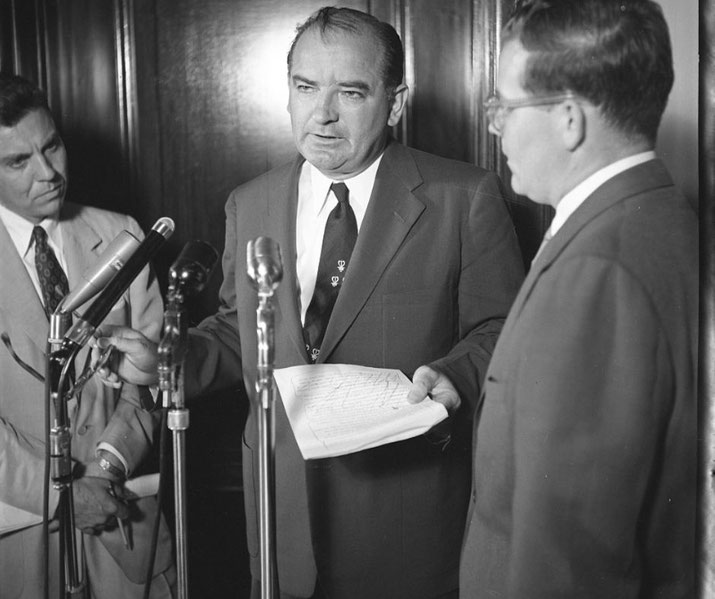 Black and white image of a man holding papers and speaking at microphones, flanked by two other suited men in what appears to be a formal press setting.