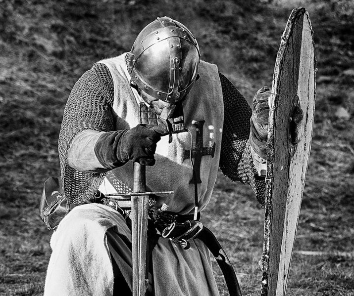 A knight in full armor kneels with head bowed, gripping a sword and shield in a solemn pose on a grassy field.