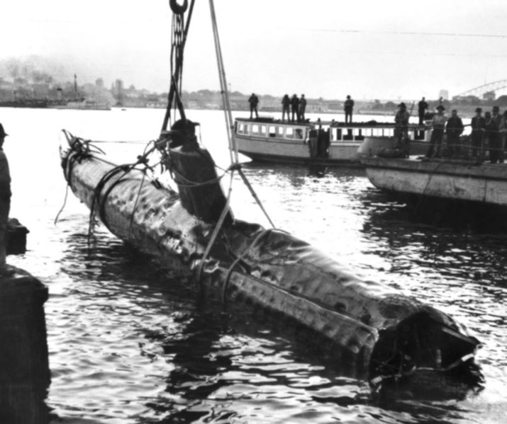 A damaged midget submarine is hoisted from the water by crane, with boats and onlookers in the background.