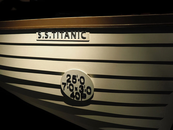 AThe side of a lifeboat from the Titanic with the label "S.S. Titanic" and capacity details "25' 0, 7' 0 x 3' 0, 40 P" prominently displayed, illuminated against a dark background.
