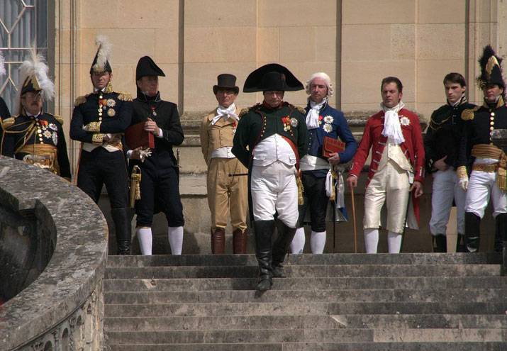 Men in period uniforms and formal attire pose on stone steps, with one figure dressed as Napoleon walking forward in a reenactment or historical event setting.