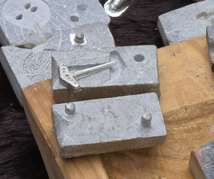 A stone mold with a partially cast silver Thor's hammer pendant, resting on a wooden surface. The intricate Nordic design suggests a historical or artisanal context for metal crafting.