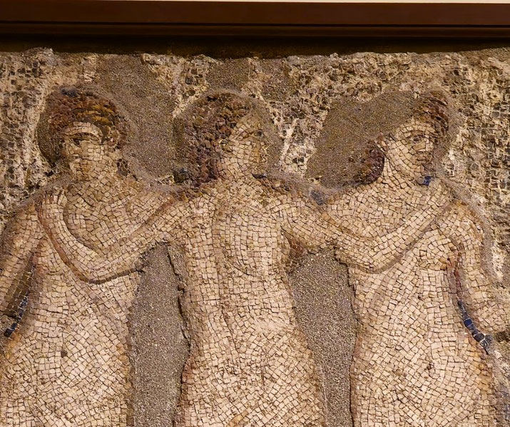 Ancient mosaic depicting three nude female figures standing arm in arm, worn with age but still retaining graceful outlines.