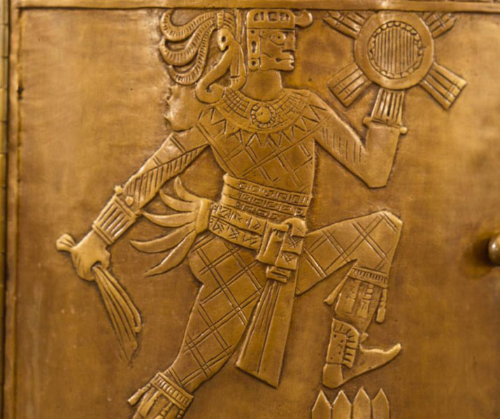A bronze relief depicting an ancient Mesoamerican warrior or deity. The figure wears elaborate armor, holds a weapon and a sun symbol, and has intricate headdress details.