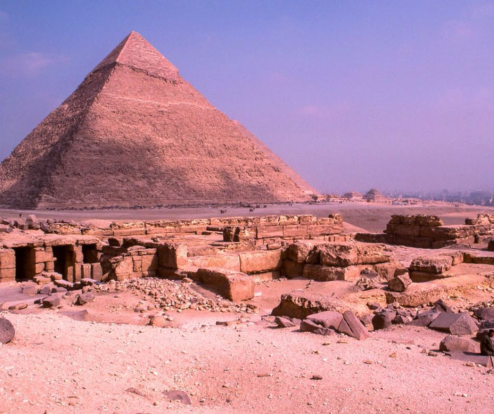 The Great Pyramid of Khufu at Giza, towering over ancient tomb ruins in the foreground. The warm desert tones contrast with the hazy blue sky, highlighting Egypt's iconic landscape.