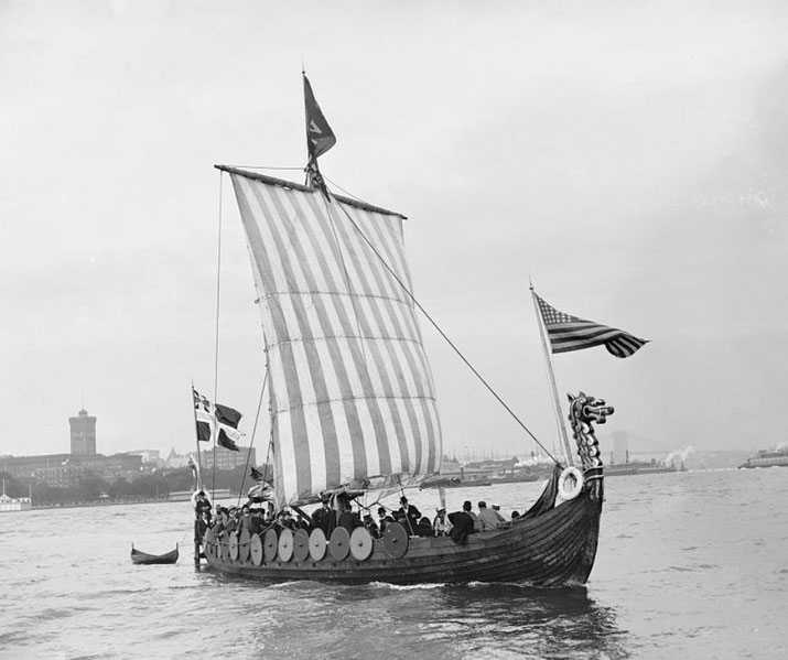 A modern reconstruction of a Viking ship with a striped sail, round shields along its sides, and a dragon-headed prow.