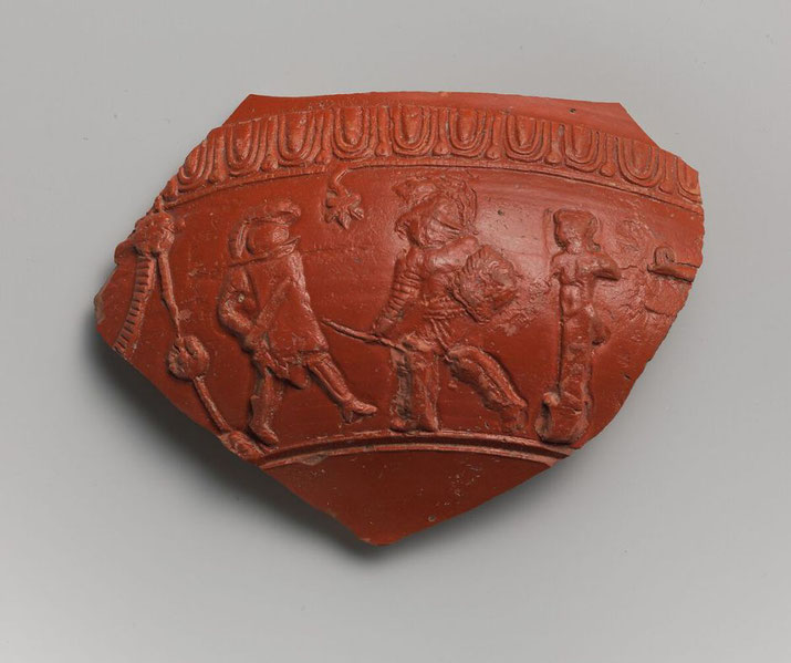 A terracotta fragment of a bowl featuring a relief of gladiators in combat. The detailed scene includes armed figures, decorative borders, and elements of Roman artistry.