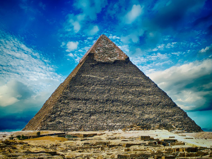 The Great Pyramid of Giza stands tall under a vibrant blue sky with scattered clouds, surrounded by ancient ruins at its base.
