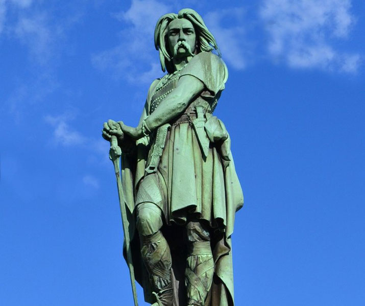Bronze statue of a warrior with long hair and mustache, gripping a sword, standing confidently against a blue sky.