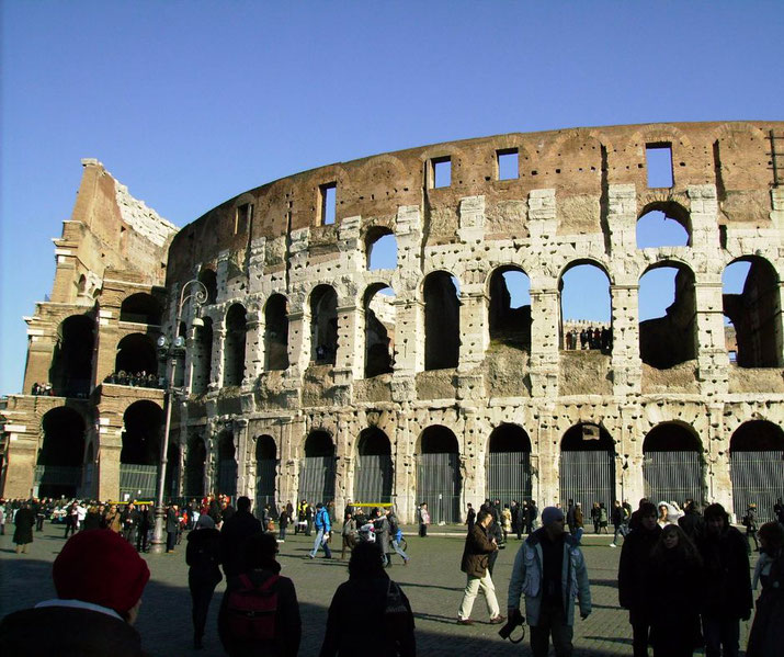 The Colosseum in Rome with its weathered arches and partial ruins. Tourists walk around the site, exploring the ancient amphitheater under a clear blue sky.