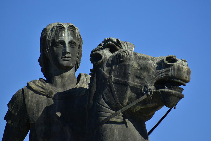 Bronze statue of a man in armor standing beside a rearing horse, both looking forward under a clear blue sky.