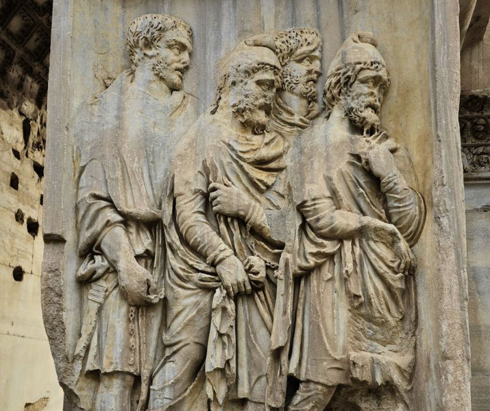 Ancient stone relief showing four bearded men in detailed robes and headwear, engaged in solemn conversation, likely depicting a historical or diplomatic scene.