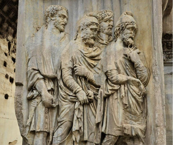 Ancient stone relief showing four bearded men in detailed robes and headwear, engaged in solemn conversation, likely depicting a historical or diplomatic scene.