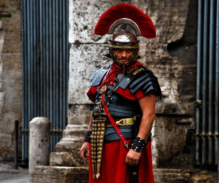 A reenactor dressed as a Roman centurion, wearing a red-crested helmet, segmented armor, and a red tunic.