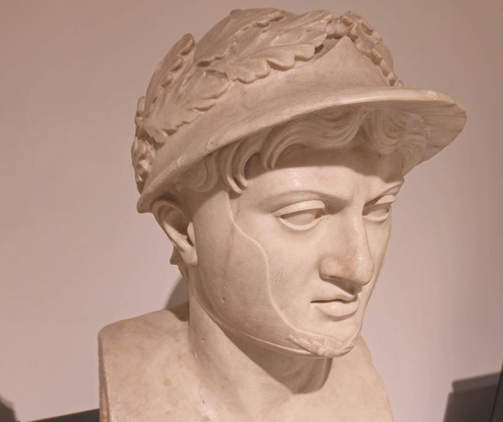 Marble bust of a helmeted man with a laurel wreath, downward gaze, and faint chin strap; detailed facial features and wavy hair under the helmet.