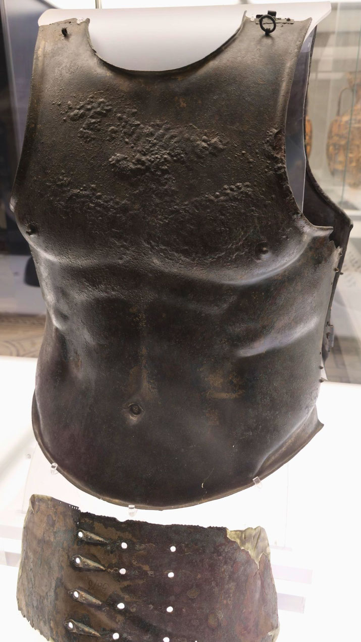 Ancient bronze cuirass with anatomical detailing, showcasing a muscular torso design and a corroded lower plate with rivet holes.