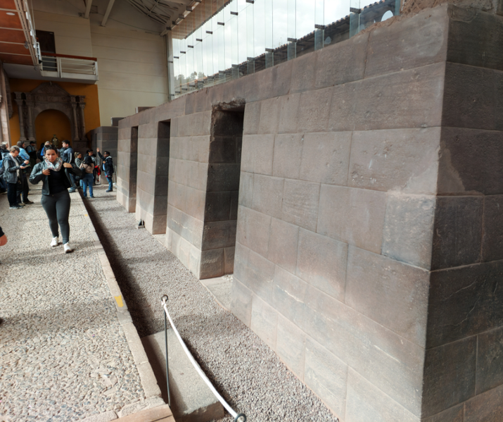 An ancient stone structure with precisely cut, fitted blocks features trapezoidal doorways. Visitors walk along a pathway beside it. 