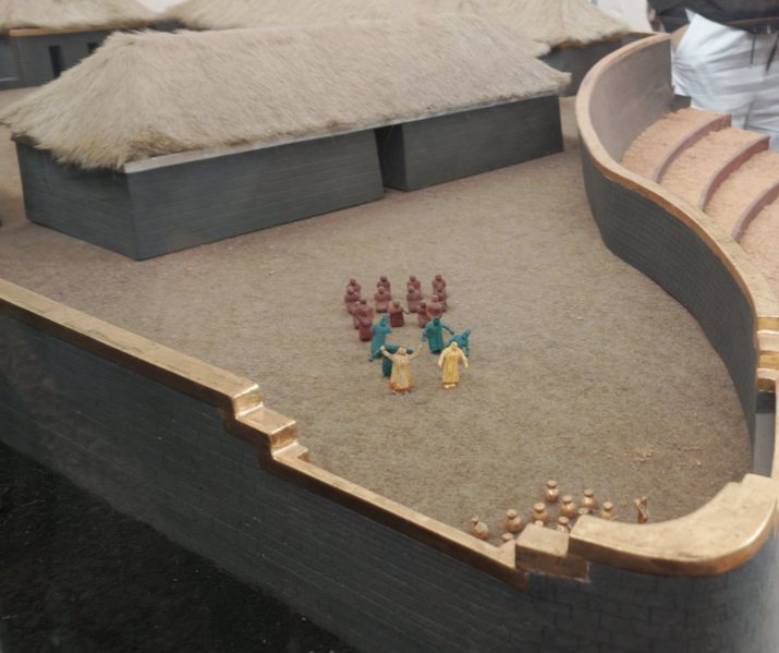A scale model of an ancient Inca site features stone structures with thatched roofs, a curved wall with gold trim, and small figurines depicting a ceremonial or social gathering in an open courtyard.