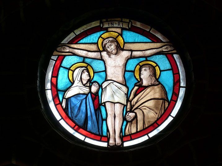 A circular stained glass window depicting the crucifixion of Jesus, with Mary and John the Apostle on either side, surrounded by vibrant red, blue, and white sections.