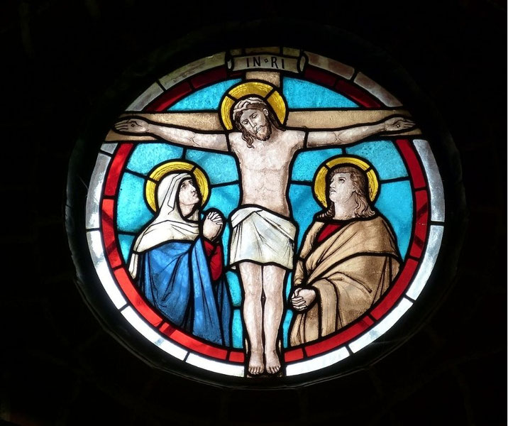 A circular stained glass window depicting the crucifixion of Jesus, with Mary and John the Apostle on either side, surrounded by vibrant red, blue, and white sections.