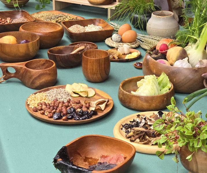 A table featuring wooden bowls and plates with nuts, seeds, dried fruits, vegetables, herbs, eggs, and rustic cups, arranged on a green cloth. The setup evokes a historical or naturalistic culinary scene.