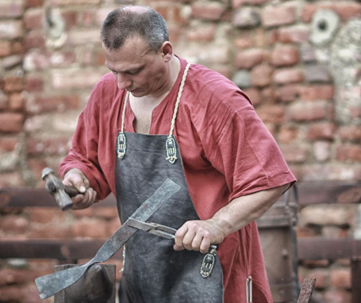 A blacksmith wearing a red tunic and leather apron hammers a piece of metal on an anvil.