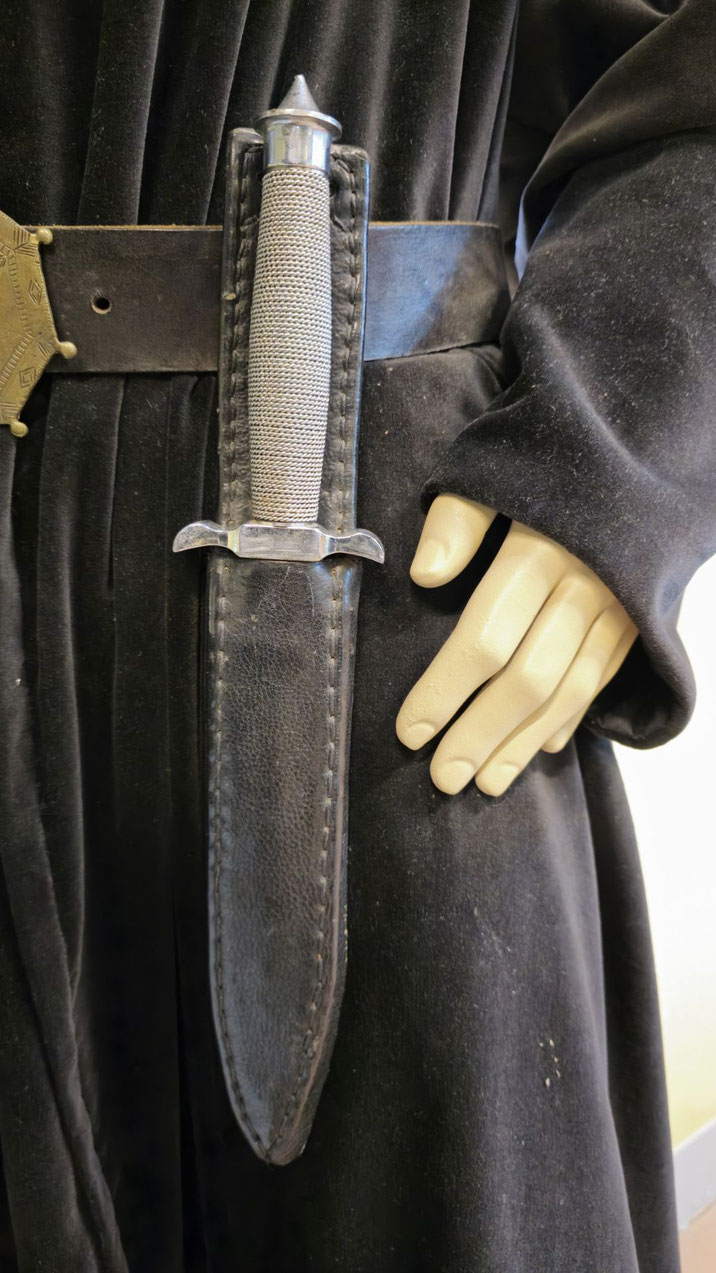 A mannequin dressed in black holds a dagger with a textured metal grip and leather sheath, attached to a wide belt.