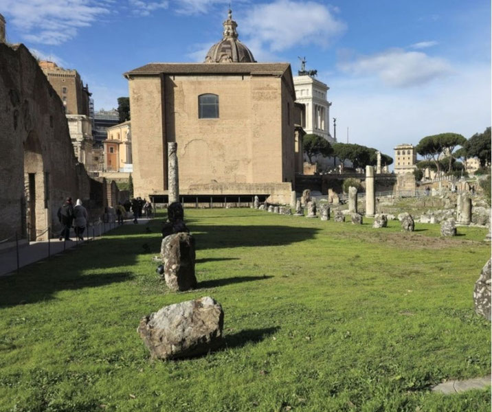 The ruins of the Basilica Aemilia in the Roman Forum. Scattered stone remnants and columns stand on a grassy area, with historic buildings and trees in the background under a blue sky.