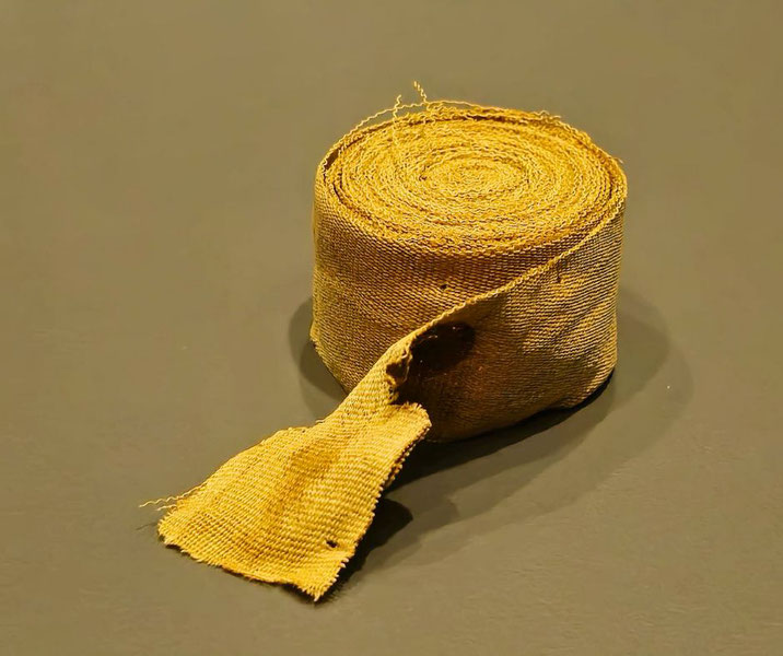 Ancient rolled linen strip used in Egyptian mummification, displayed on a dark surface with frayed end visible.