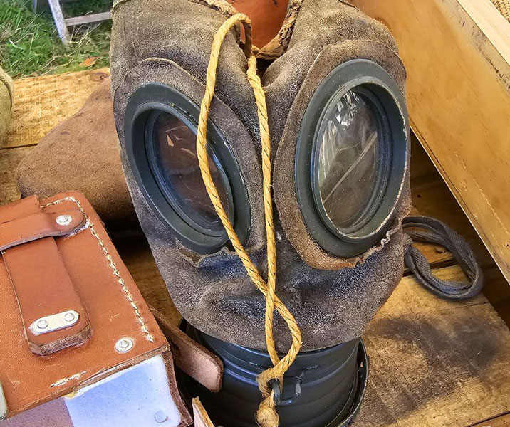 A worn World War I gas mask with large round eye lenses and a rope tie rests on a wooden surface beside leather gear.