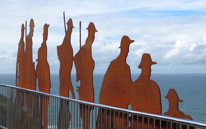 Rust-colored metal soldier silhouettes with engraved names stand on a seaside memorial platform, honoring fallen troops against an ocean backdrop.