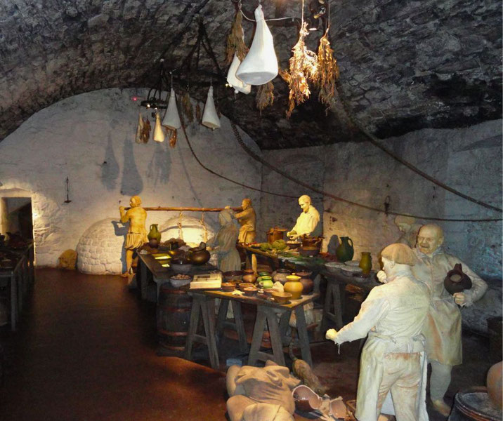  A historical kitchen scene in a stone cellar with life-sized statues depicting cooks and workers preparing food. The setting includes hanging herbs, pottery, tables, barrels, and a rustic oven.