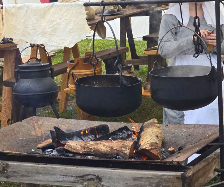 An outdoor cooking setup with iron cauldrons hanging over a wood fire, surrounded by rustic wooden furniture and a person in period attire, evoking a historical or medieval cooking scene.