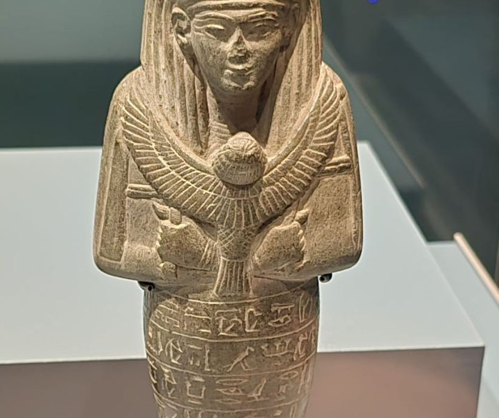 Stone funerary figurine with crossed arms, winged chest emblem, and hieroglyphs on the lower half, displayed in a museum setting.