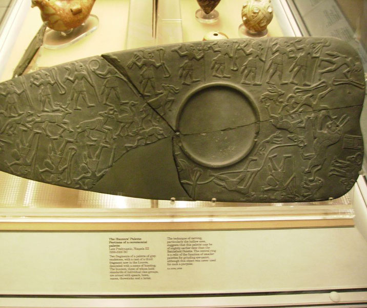 Ancient Egyptian Hunters' Palette fragment, carved with figures and animals in ceremonial scenes, displayed in a museum case.