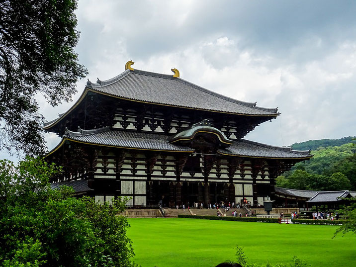 A large wooden temple with curved roofs and golden ornaments stands beside a wide green lawn under a cloudy sky, with visitors gathered near the entrance.