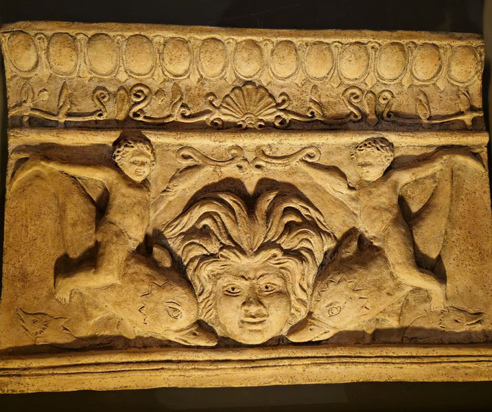 Stone relief showing a central face with flowing hair, flanked by two children riding dolphins, with decorative patterns along the top.