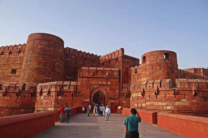 Tourists walk toward the massive red sandstone walls and arched entrance of a historic fort under a clear sky.