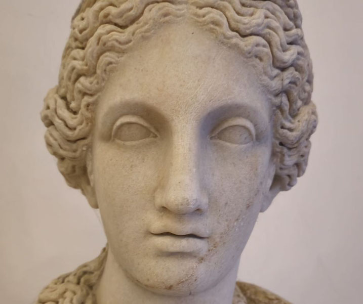 Close-up of a marble statue's face with wavy hair and blank, stylized eyes, showcasing classical beauty and serene expression.