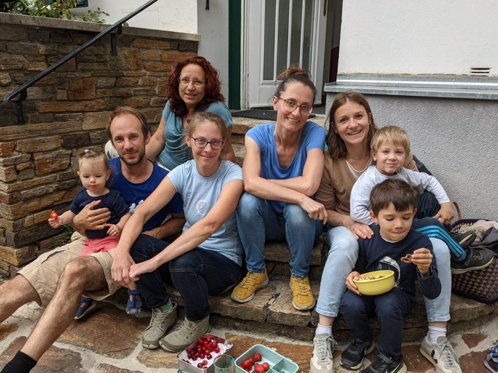 Das Team der Praxis im Jahr 2022. Die Personen sitzen auf der Stiege vorm Einang zur Praxis. Es sind 3 Kinder der Therapeuten anwesend, sie essen gerade Obst. Das Bild wirkt sehr persönlich und familiär.
