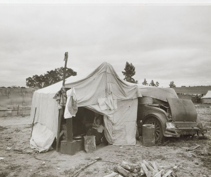 A makeshift tent made from cloth and an old car serves as shelter for a family during the Great Depression, with items scattered outside on the dirt.