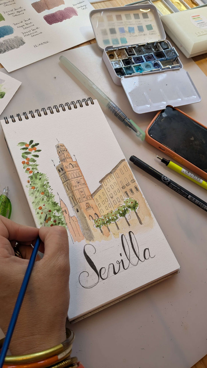Une peinture à l'aquarelle montre des scènes de Séville et de Grenade. Des palettes de couleurs, des pinceaux, et des crayons sont visibles sur la table. Les illustrations incluent des bâtiments historiques et des paysages.
