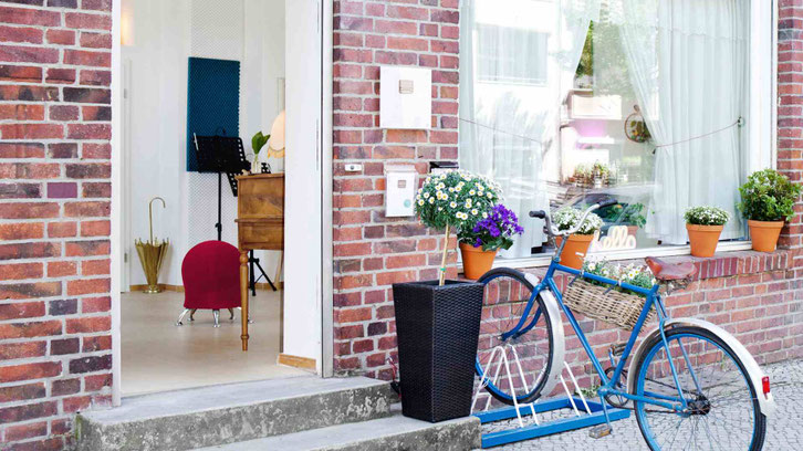 Eingang des STIMME+ Studios in Berlin mit geöffneter Tür, Blick in einen hellen Raum mit Klavier, Sitzmöbel und Pflanzen, davor ein Fahrrad und Blumentöpfe.