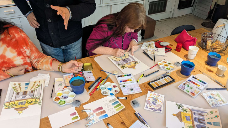 Des personnes peignent à l'aquarelle autour d'une table avec des pinceaux, des crayons, des palettes de couleurs, et des tubes de peinture. Plusieurs illustrations de bâtiments et de paysages sont visibles, ainsi que des feuilles avec des échantillons