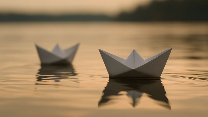 Zwei weiße Papierboote treiben auf ruhigem Wasser in warmem Abendlicht. Das vordere Boot ist scharf fokussiert, das hintere verschwommen im Hintergrund. Die sanften Wellen spiegeln das goldene Licht, die Szene wirkt still, poetisch und kontemplativ.