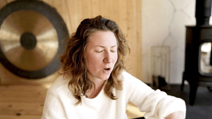 Marie Rodewald sitzt mit geschlossenen Augen in einem warmen Raum mit Gong im Hintergrund und ist im Klang oder in stimmlicher Arbeit vertieft.