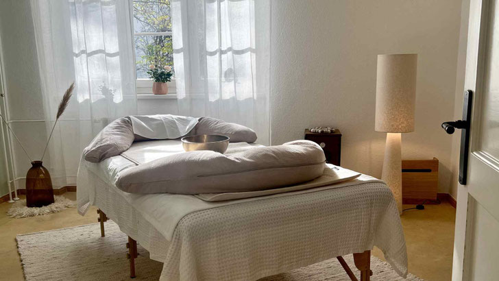 Heller, ruhiger Raum mit Liege, Kissen und Klangschale, weiches Licht durch ein Fenster mit Vorhängen, minimal und warm eingerichtet.