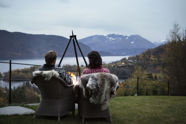 Zwei Personen von hinten vor einem Lagerfeuer in mit Fell belegten Korbstühlen mit Ausblick auf den Fjord