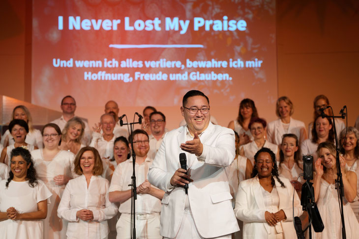 Hanjo in weiß vor dem weiß gekleideten Chor lächelt und deutet Richtung Kamera